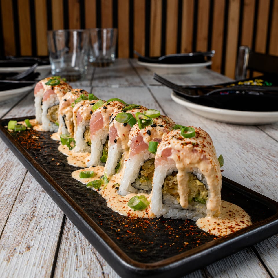 Sushi rolls topped with tuna, creamy sauce, green onions, and spices on a black plate.
