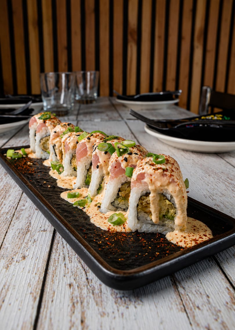 Sushi rolls topped with tuna, creamy sauce, green onions, and spices on a black plate.