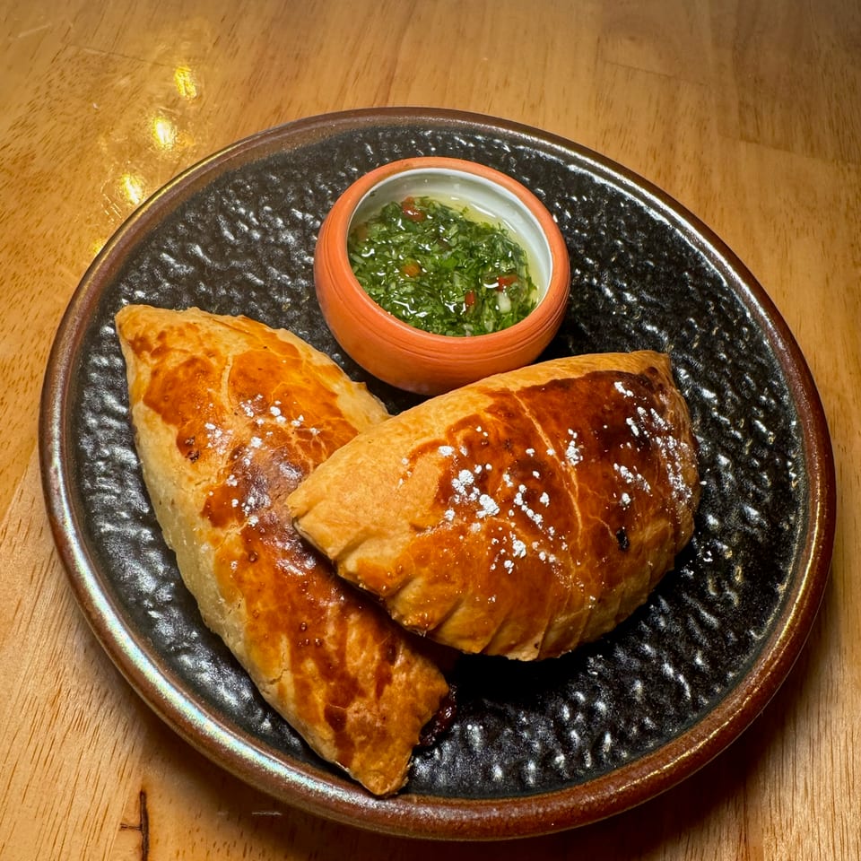 Two golden-brown empanadas served on a black ceramic plate with a small bowl of green herb sauce.