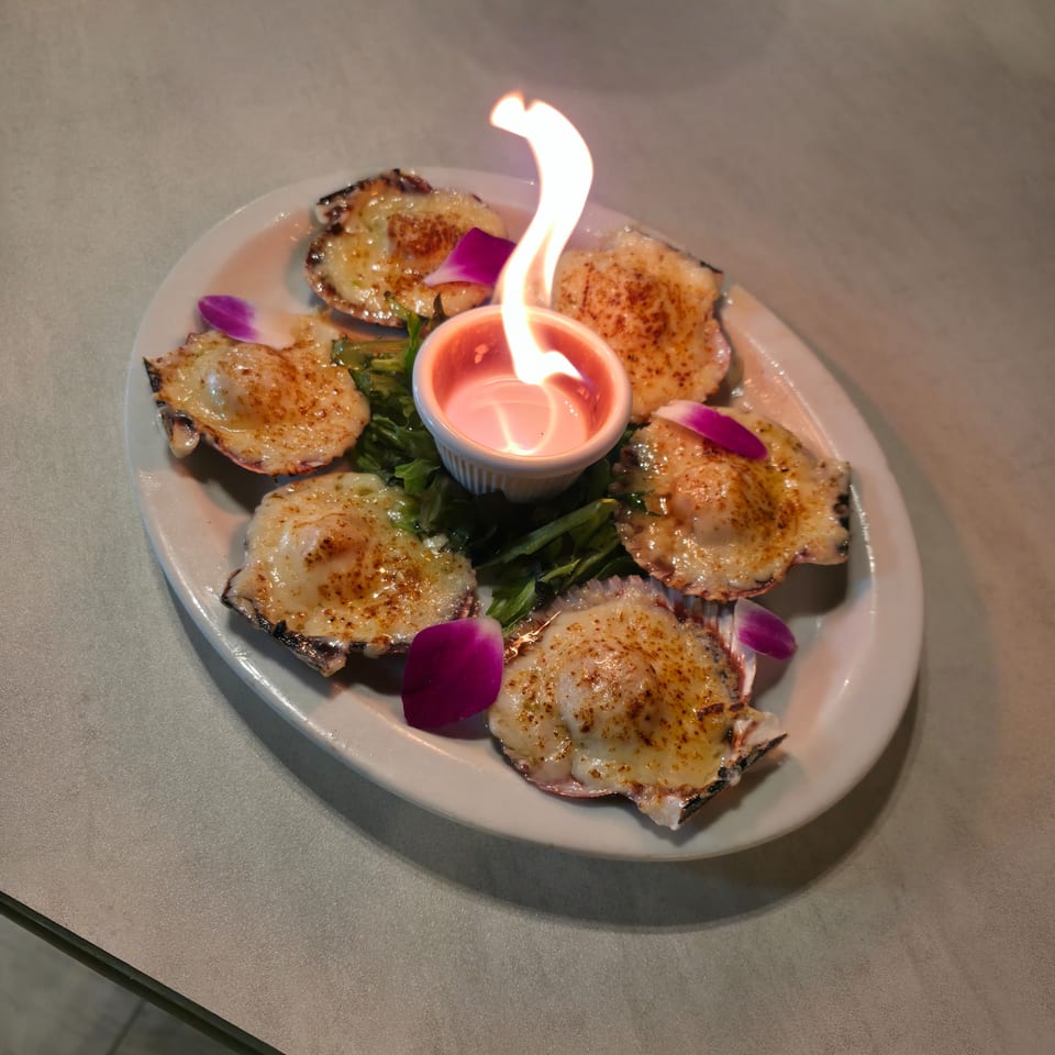 Broiled scallops topped with melted cheese surround a flaming ramekin of sauce, garnished with greens and purple flower petals.