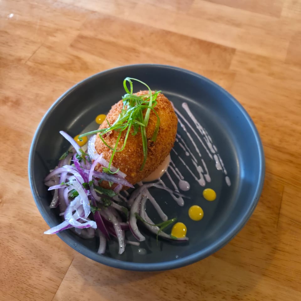 Crispy breaded croquette topped with green herbs, served with red onion salad and colorful sauce drizzles on a dark plate.