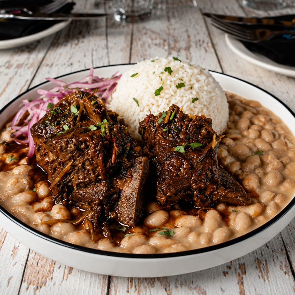 Braised beef served with white rice, creamy beans, pickled onions, and fresh herbs on a rustic wooden table.