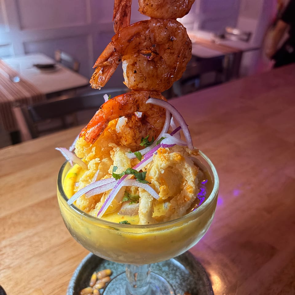 Cocktail glass topped with grilled shrimp, crispy fried seafood, red onion, herbs, and yellow sauce in a dimly lit restaurant.