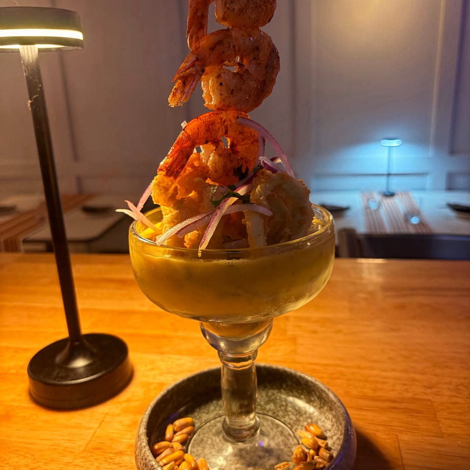 Golden cocktail in a stemmed glass garnished with stacked grilled shrimp, vegetables, and edible flowers on a restaurant table.