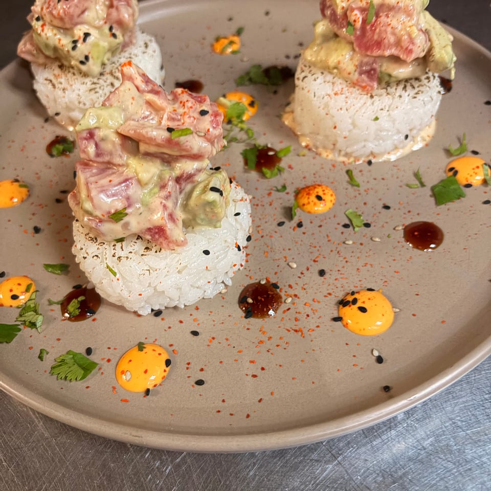 Three sushi rice towers topped with creamy salmon, tuna, and avocado, garnished with sesame seeds, herbs, spicy mayo, and sauces.