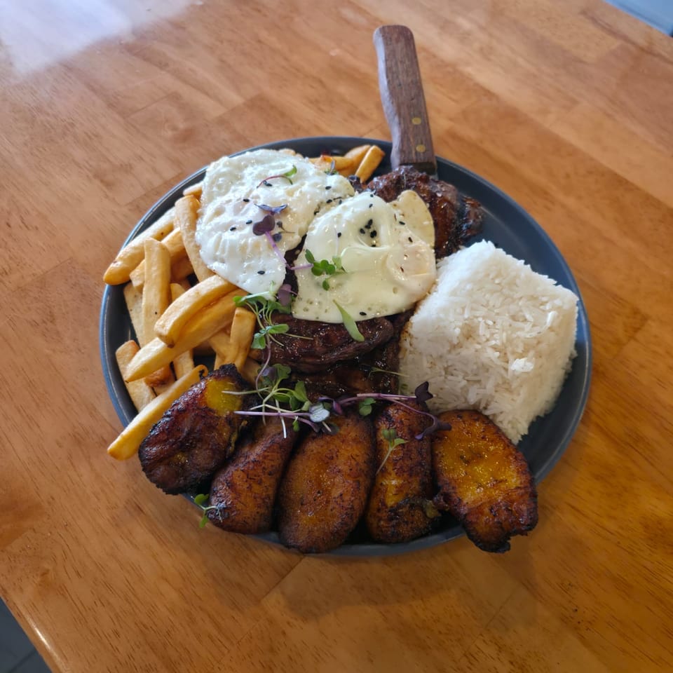 Grilled steak topped with fried eggs, served with rice, fries, plantains, and microgreens on a black plate.