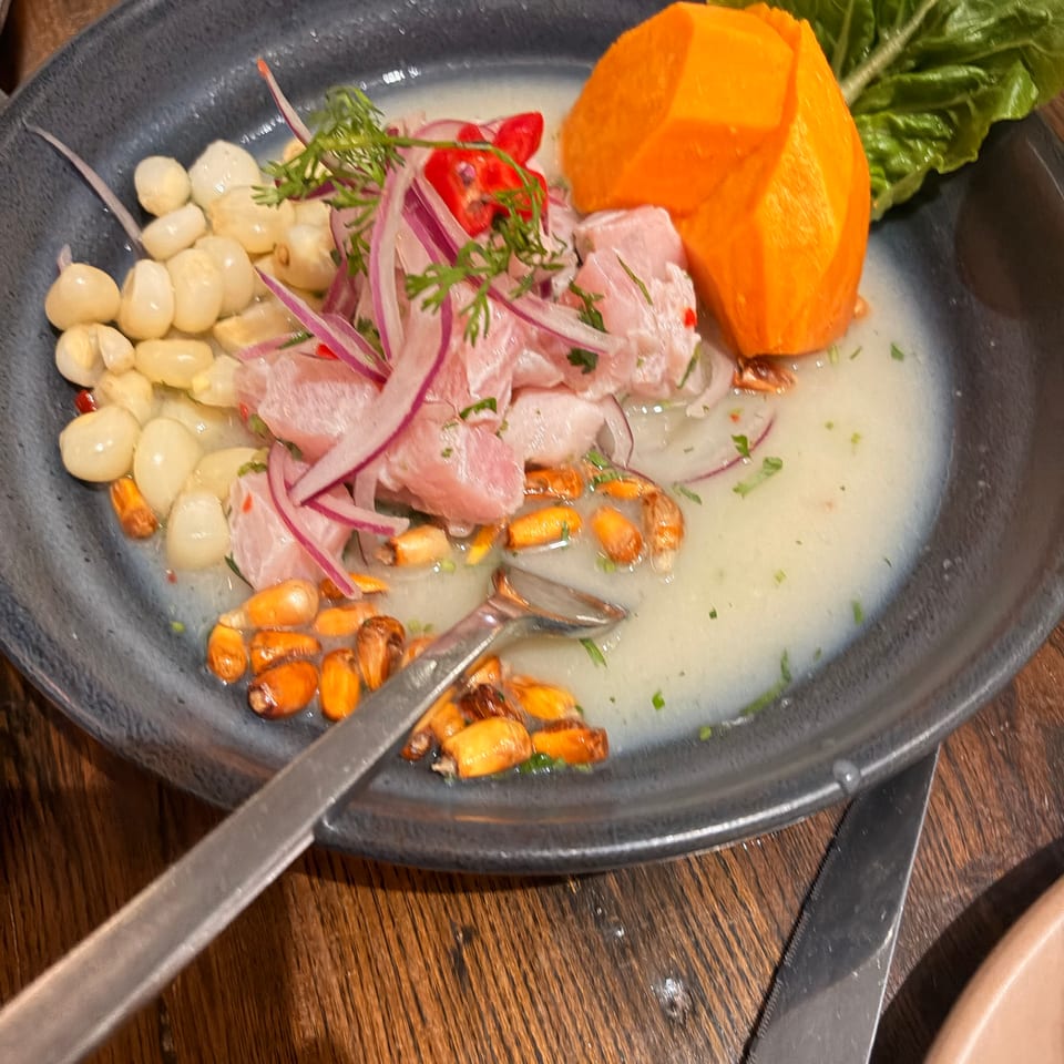 Ceviche with white fish, red onion, corn, roasted corn kernels, herbs, lettuce, and an orange garnish in a dark bowl.