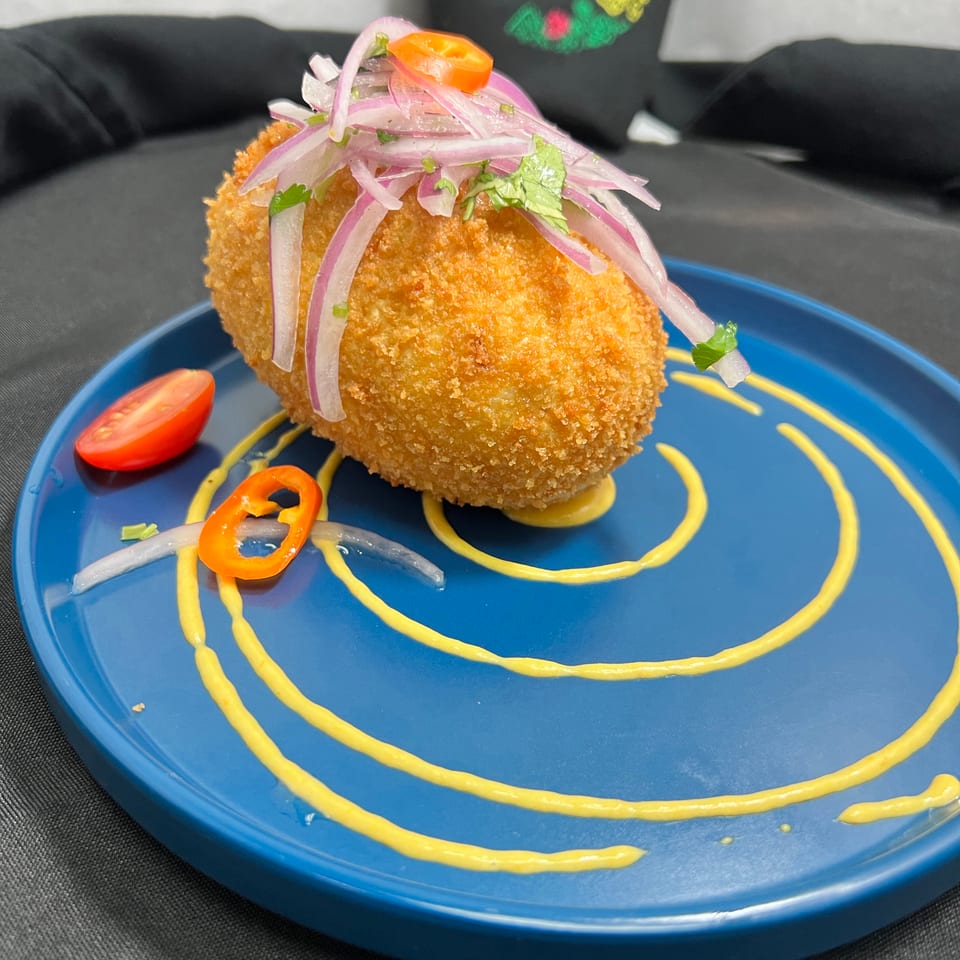 Golden-brown breaded dish topped with pickled red onions, herbs, and peppers on a blue plate drizzled with yellow sauce.
