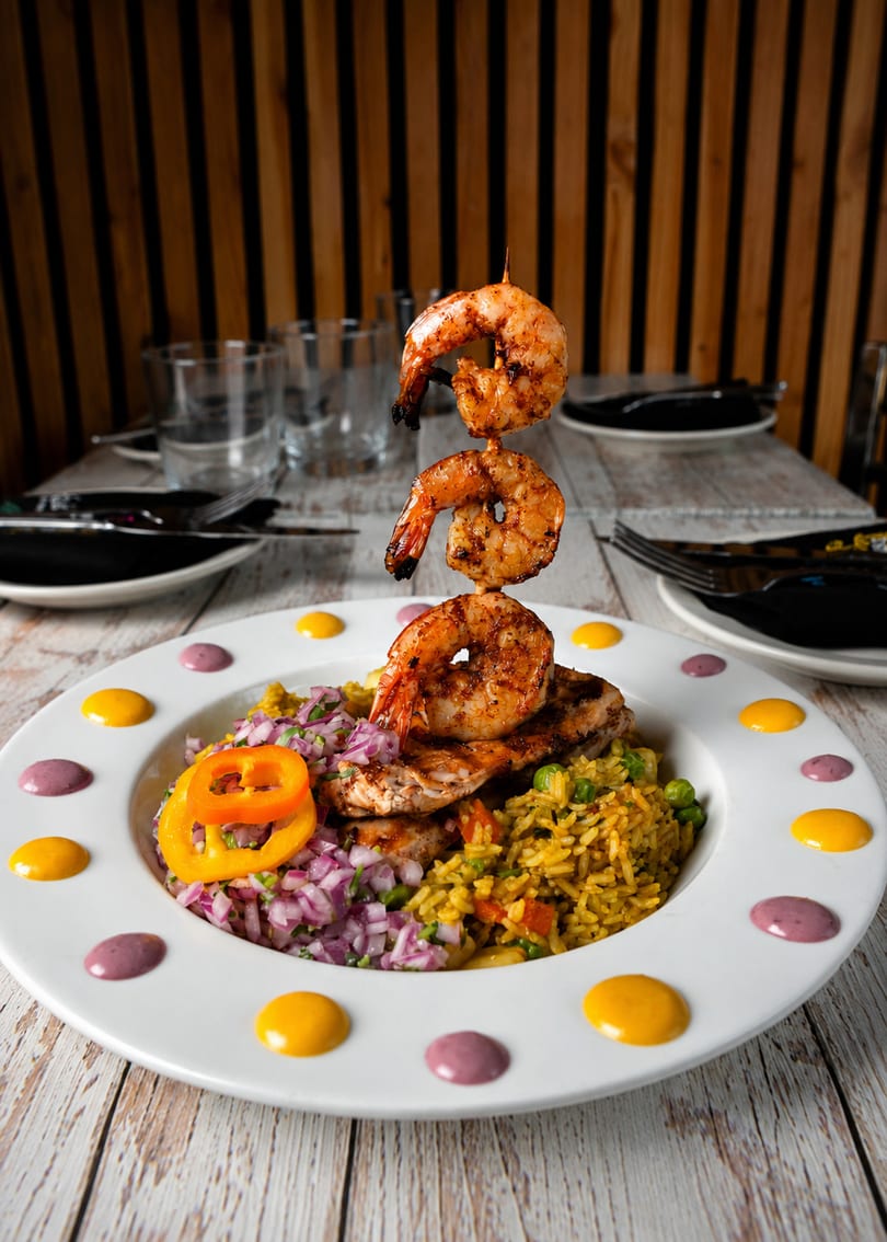 Grilled shrimp skewer over chicken, yellow rice, and red onion relish on a white plate.