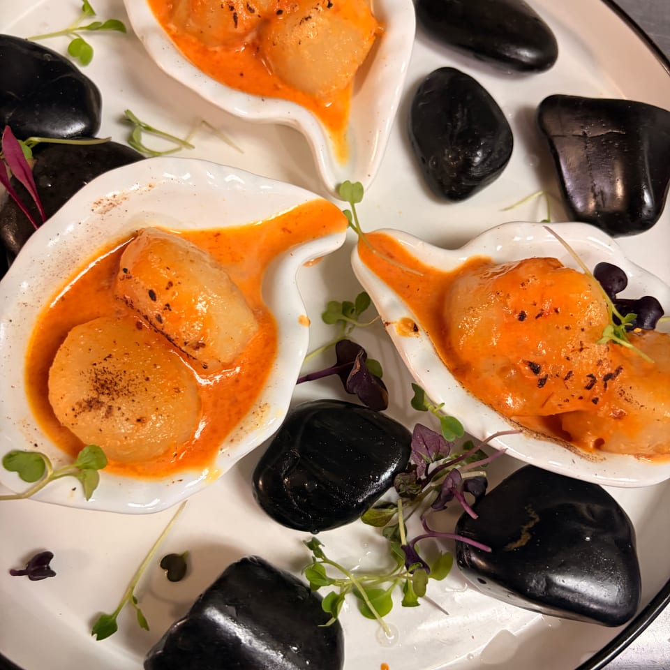 Three white dishes hold seared scallops in bright orange sauce, garnished with microgreens and surrounded by black decorative stones.