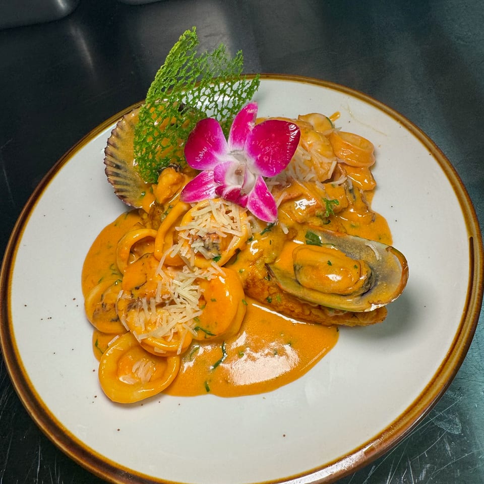 Plate of seafood pasta in creamy orange sauce, topped with mussels, shredded cheese, herbs, and a purple orchid garnish.