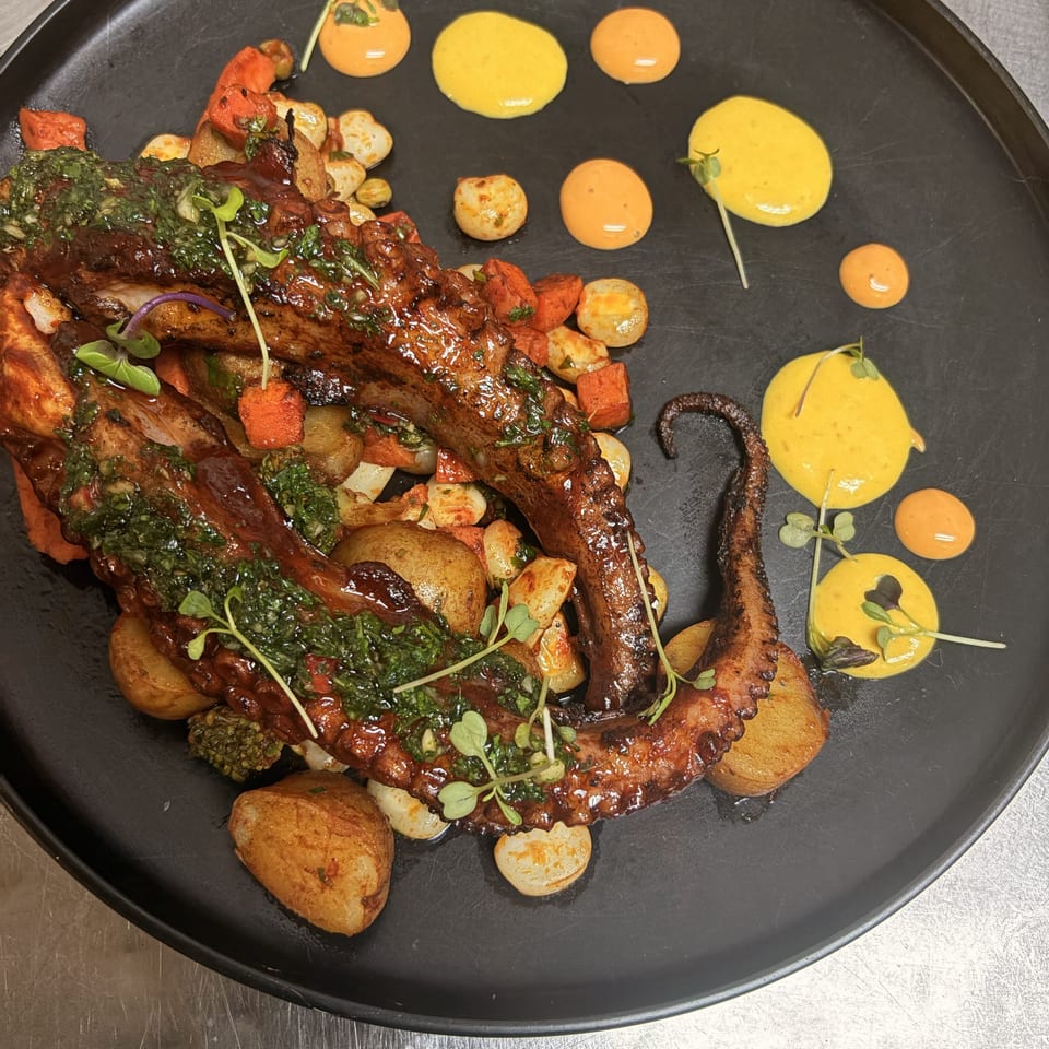 Glazed grilled octopus tentacles with chimichurri, roasted potatoes, vegetables, microgreens, and colorful sauces on a black platter.