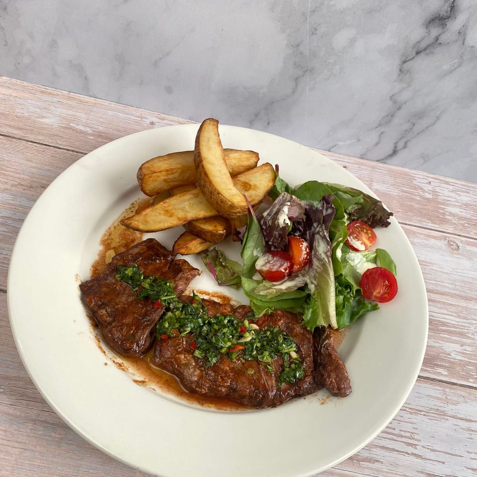 Grilled steak topped with green herb sauce, roasted potato wedges, and mixed-leaf salad with cherry tomatoes on a plate.