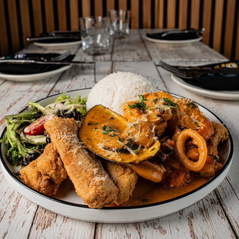 Large platter of crispy breaded chicken, seafood in creamy sauce, white rice, and salad on a restaurant table.
