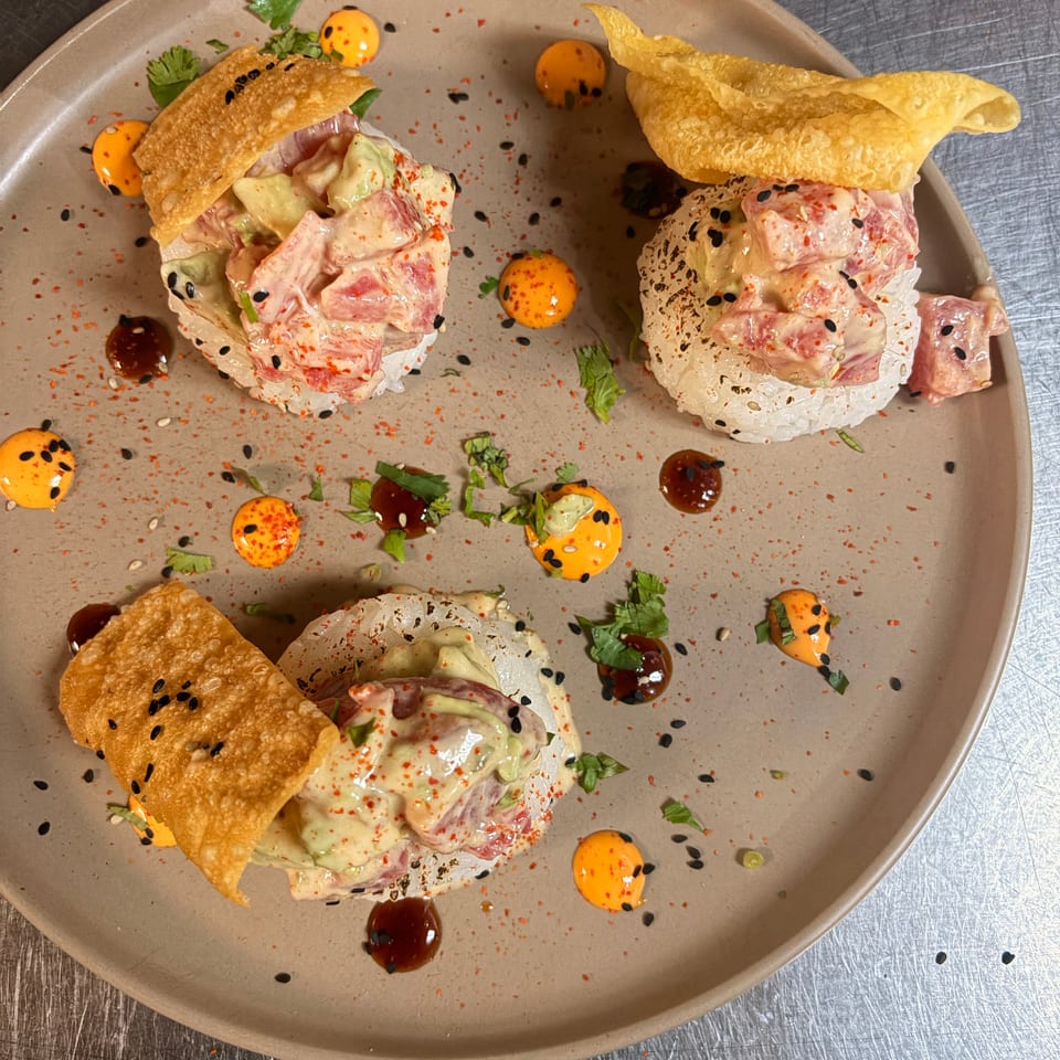 Three crispy taco-style appetizers topped with creamy diced seafood, avocado, herbs, black sesame seeds, and colorful sauces on a plate.
