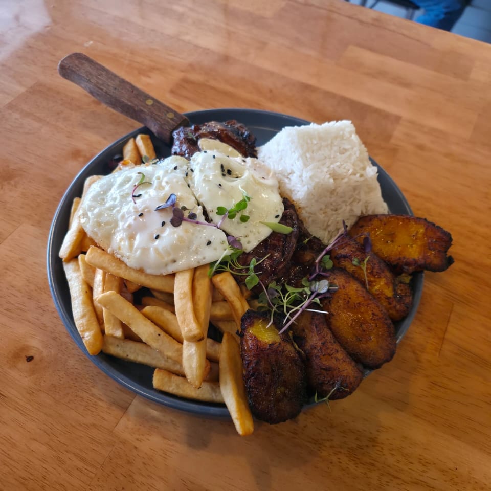 Plated grilled steak topped with fried eggs and sauce, served with fries, white rice, grilled plantains, and microgreens.