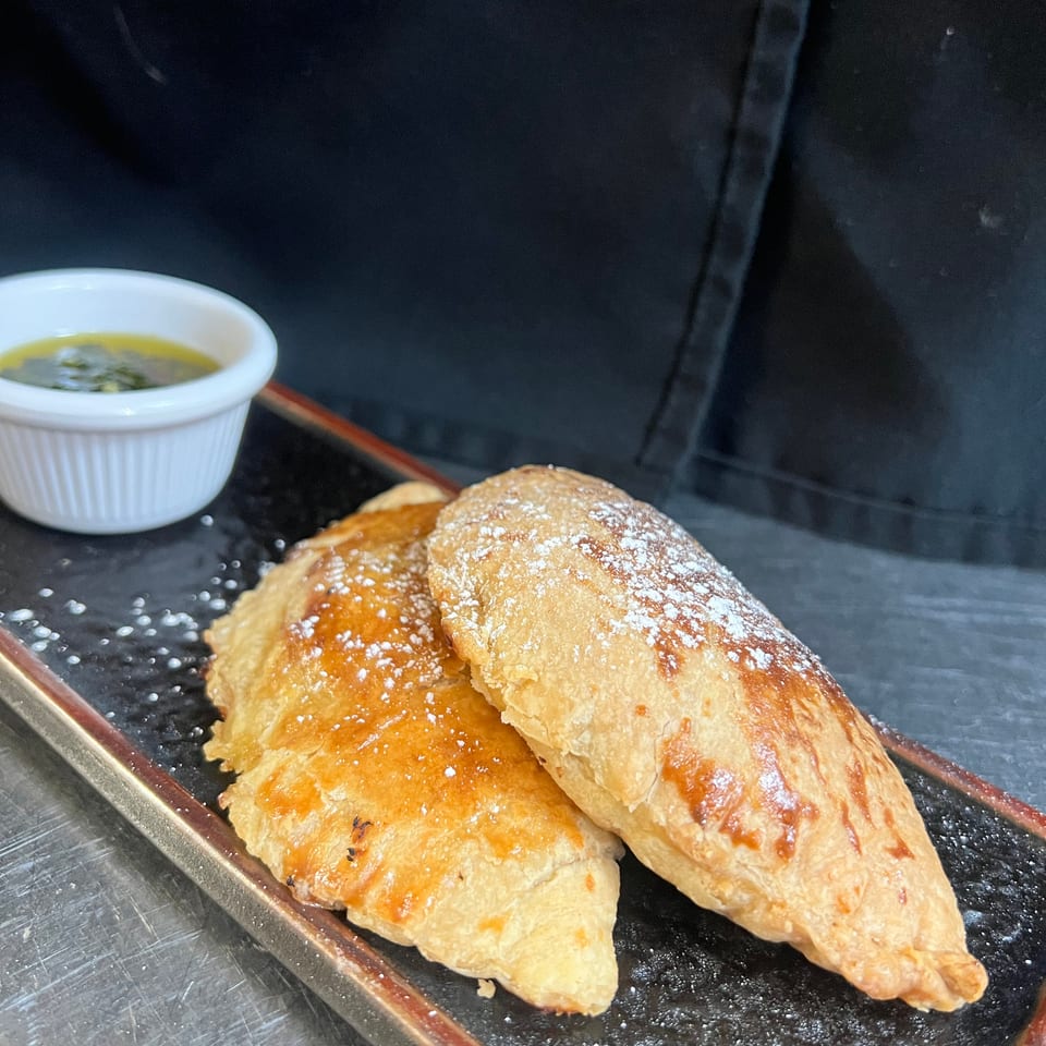 Golden flaky pastries dusted with powdered sugar sit on a black serving tray beside a small bowl of dipping sauce.