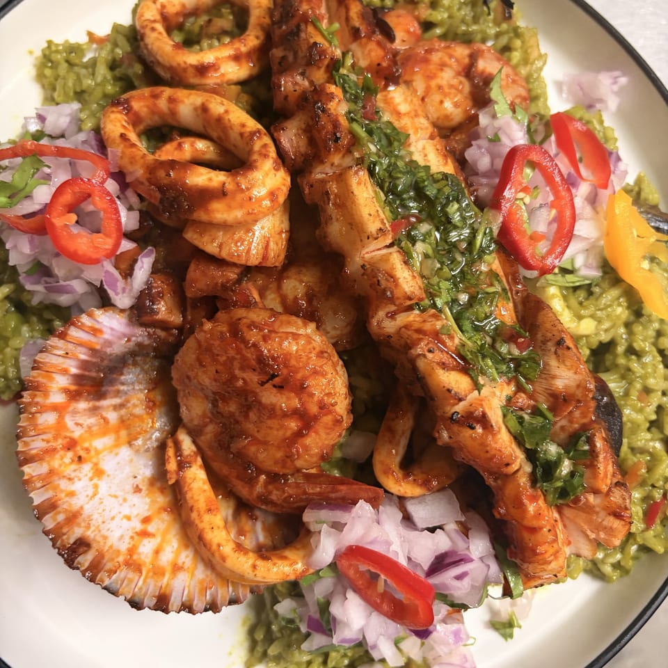 Grilled seafood medley with squid, shrimp, octopus, and scallops over green rice, garnished with red onion and chili.