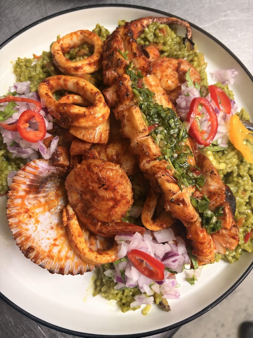 Grilled seafood medley with squid, shrimp, octopus, and scallops over green rice, garnished with red onion and chili.