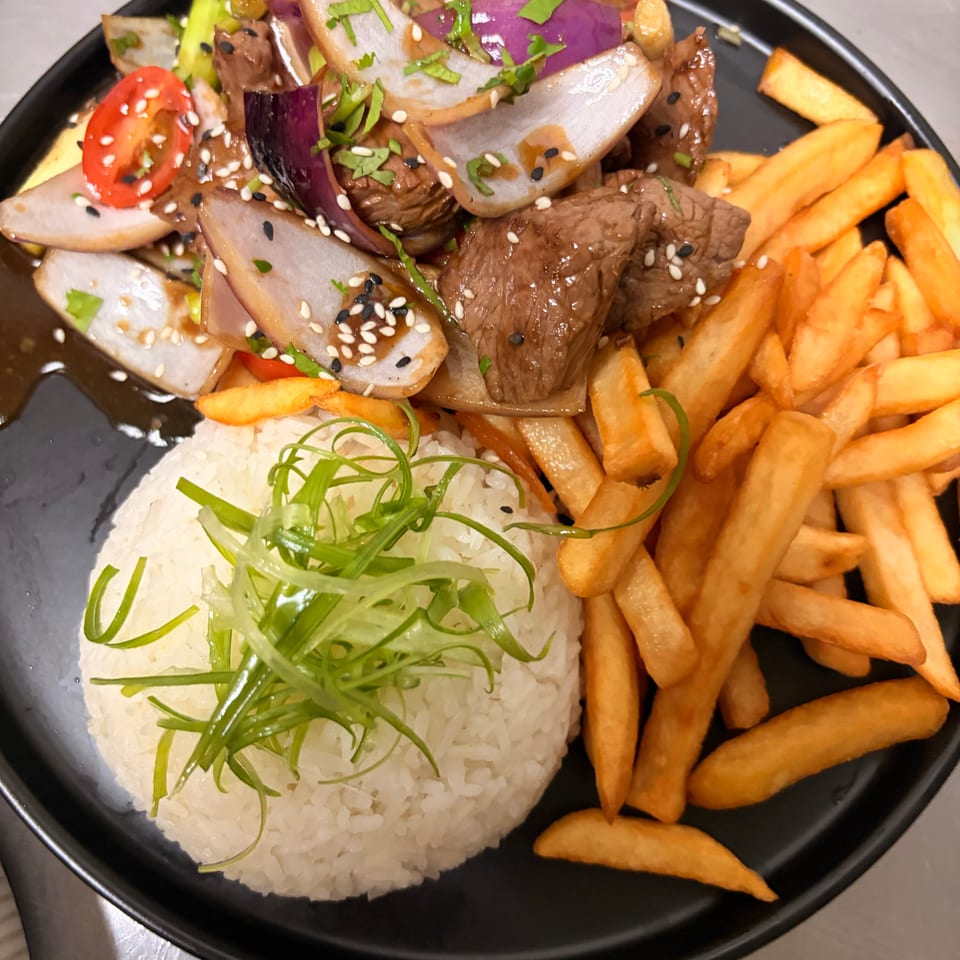 Black plate of glazed beef with onions, tomatoes, vegetables, white rice, and golden fries, garnished with sesame seeds and herbs.