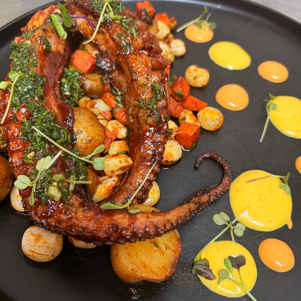 Grilled octopus tentacles with roasted potatoes, vegetables, chimichurri, microgreens, and colorful sauces on a black plate.